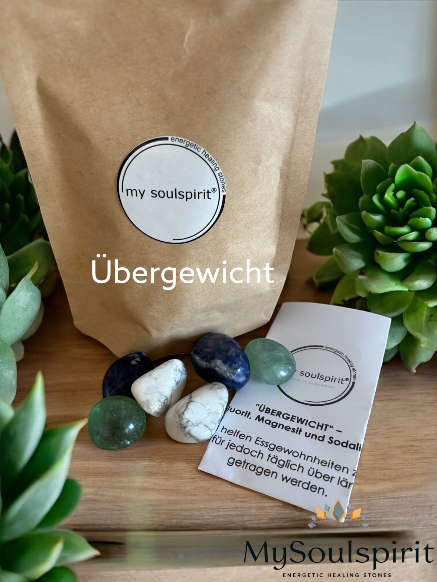 Das Heilstein-Set übergewicht besteht aus 6 Trommelsteinen von 3 Sorten die einer modernen Holzplatte vor der braunen Verpackung liegen. im Hintergrund grüne Pflanzen als Dekoration.