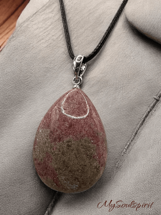 Eine Nahaufnahme eines tropfenförmigen Rhodonit-Anhängers mit silberner Fassung an einem schwarzen Lederband, der auf einem grauen, leicht strukturierten Stoff-Untergrund liegt.